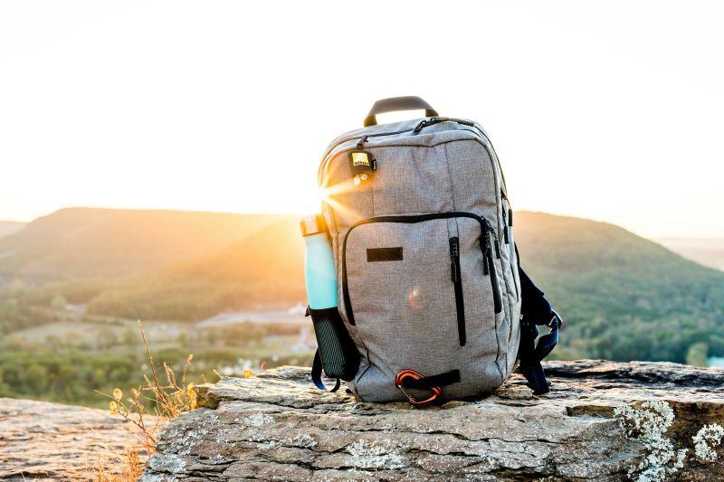 A grey hiking backpack with a reusable water bottle in its side pocket on a rock at sunset