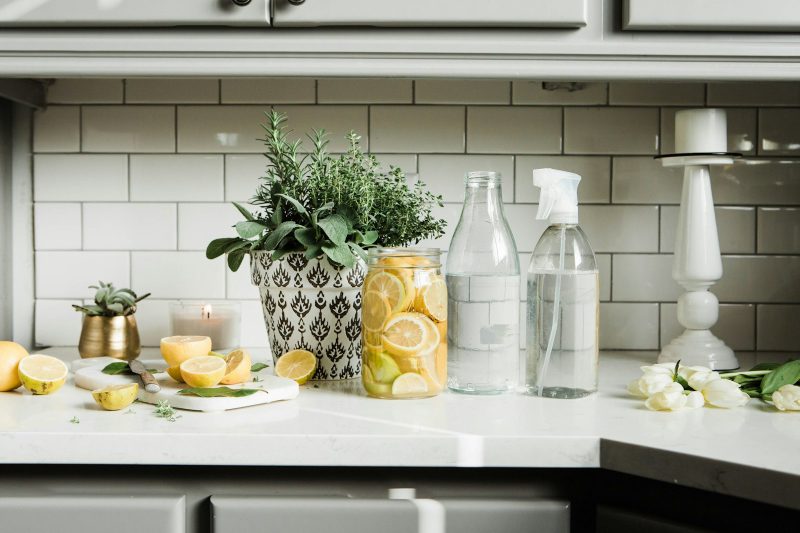 Clear glass bottles including a spray bottle on a white kitchen counter with lemons and herbs