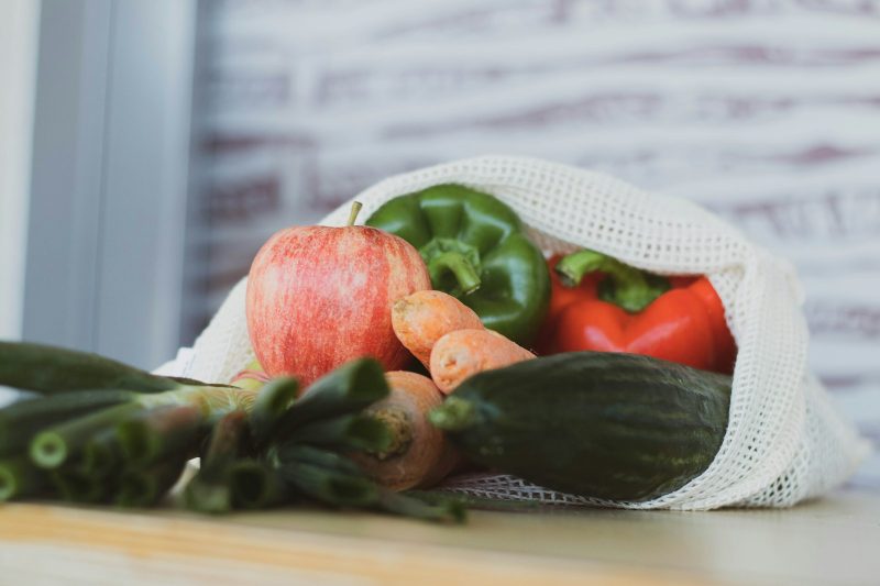 A cotton mesh produce bag filled with peppers, apples and cucumbers on a kitchen counter