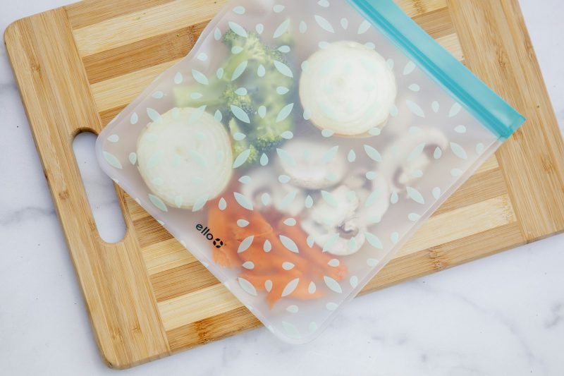 A reusable zip-seal food bag filled with sliced vegetables on a wooden chopping board