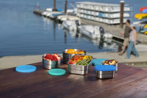 Round stainless steel food containers with coloured silicone lids on an outdoor wooden table